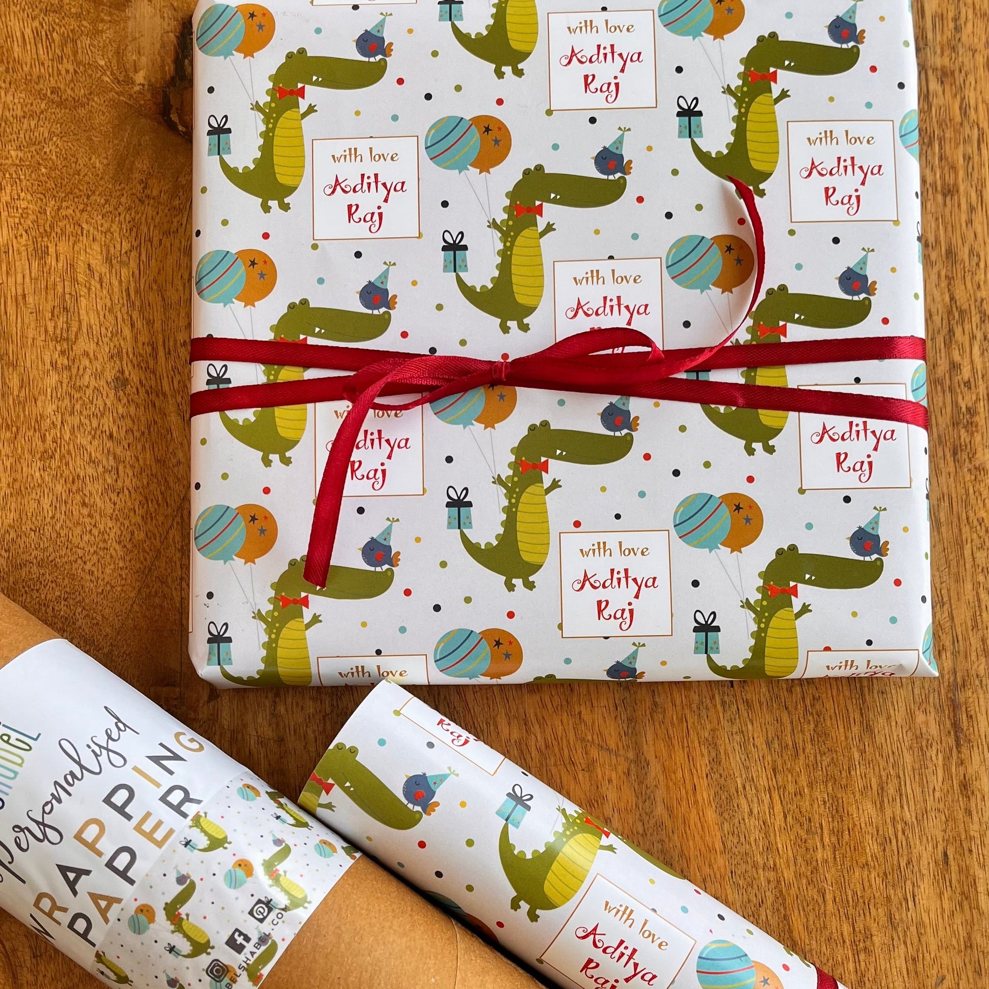 A gift set of wrapping paper and a brown kraft paper tube on a wooden table. The wrapping paper is white with a colorful cartoon crocodile pattern and a red ribbon tied around the main package.