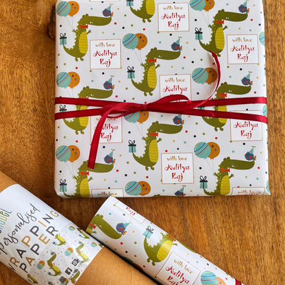 A gift set of wrapping paper and a brown kraft paper tube on a wooden table. The wrapping paper is white with a colorful cartoon crocodile pattern and a red ribbon tied around the main package.