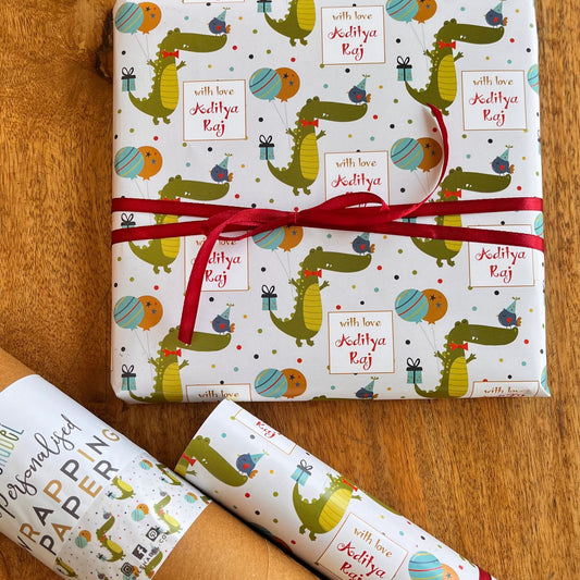 A gift set of wrapping paper and a brown kraft paper tube on a wooden table. The wrapping paper is white with a colorful cartoon crocodile pattern and a red ribbon tied around the main package.