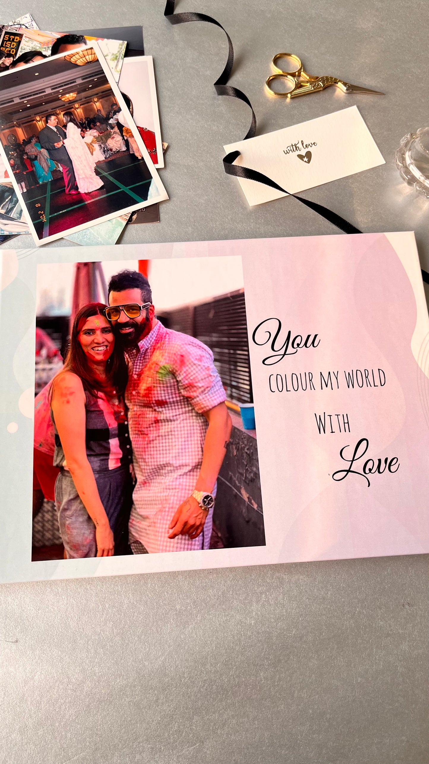 A custom printed photo book with a pink and white background, featuring a couple's photo and the text 'You COLOUR MY WORLD WITH Love'. The book is styled on a gray surface with gold scissors, a black ribbon, and other printed photos.