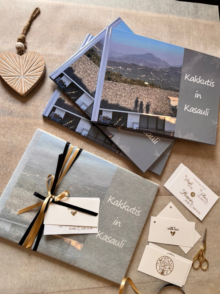 A styled flat-lay of printed wedding or event stationery on a beige fabric surface, including a wrapped gift box, a stack of photo cards or brochures, small white cards with gold and black text, scissors, and a heart-shaped ornament. 