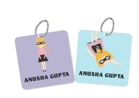 Two square metal bag tags with a character illustration and the name 'Anusha Gupa' printed on them, one tag is pink and the other is blue.