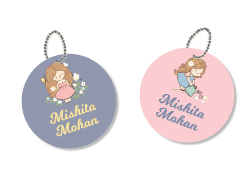 Two round metal bag tags with chain attachments. One has a gray background with a floral design and the name 'Misha', while the other has a pink background with a similar floral design and the name 'Mohan'.