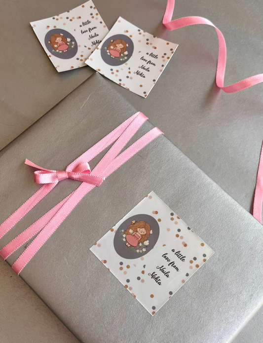 A wrapped gift box in gray paper with a pink ribbon and a decorative paper label, placed on a gray surface with two similar labels nearby.