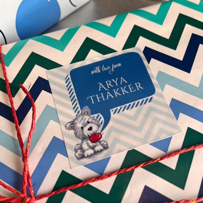 A close-up of a personalized gift tag with a blue speech-bubble design reading 'with love from ARYA THAKKER' and a small dog illustration, placed on top of chevron-patterned wrapping paper with red string.