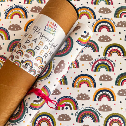 A roll of wrapping paper with a colorful rainbow and cloud pattern, partially unrolled over a brown kraft cardboard tube. The pattern features repeating rainbows in red, orange, yellow, green, blue, and purple on a white background, with brown clouds and small stars.