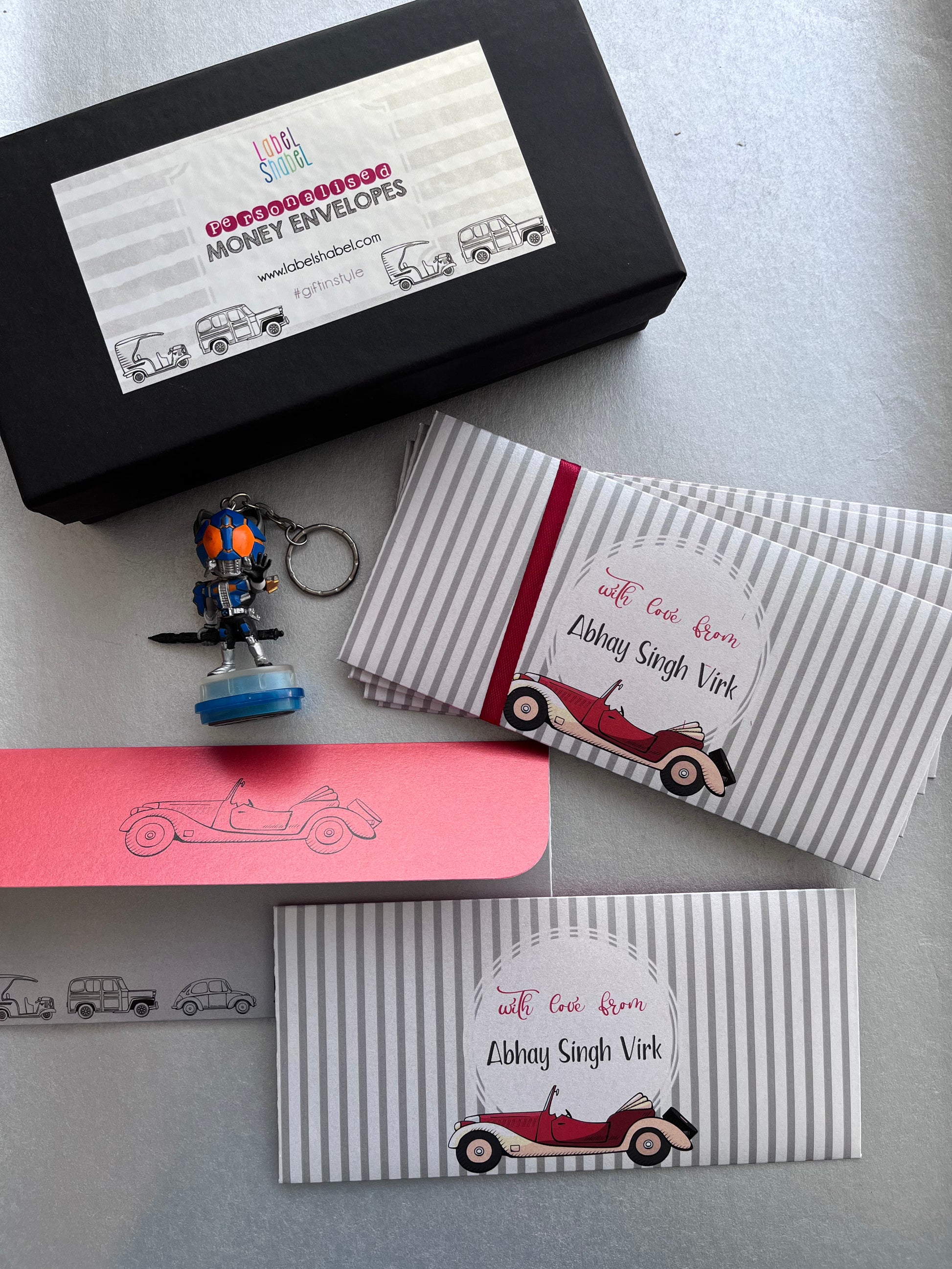 A gift set of personalized striped envelopes, a black branded box, a pink card, and a small robot keychain on a light gray surface. The envelopes are white and gray striped with red ribbon bands and a vintage car illustration, featuring the text 'with Gopi from Abhay Singh Vird'.