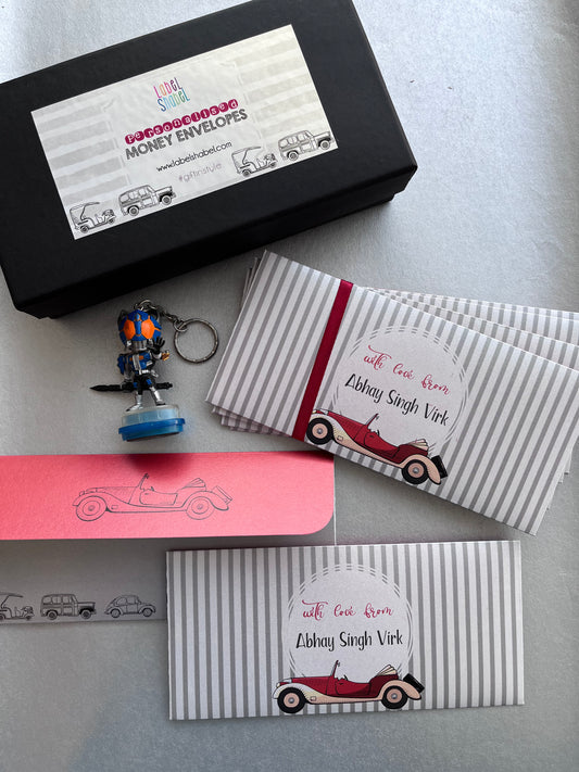 A gift set of personalized striped envelopes, a black branded box, a pink card, and a small robot keychain on a light gray surface. The envelopes are white and gray striped with red ribbon bands and a vintage car illustration, featuring the text 'with Gopi from Abhay Singh Vird'.