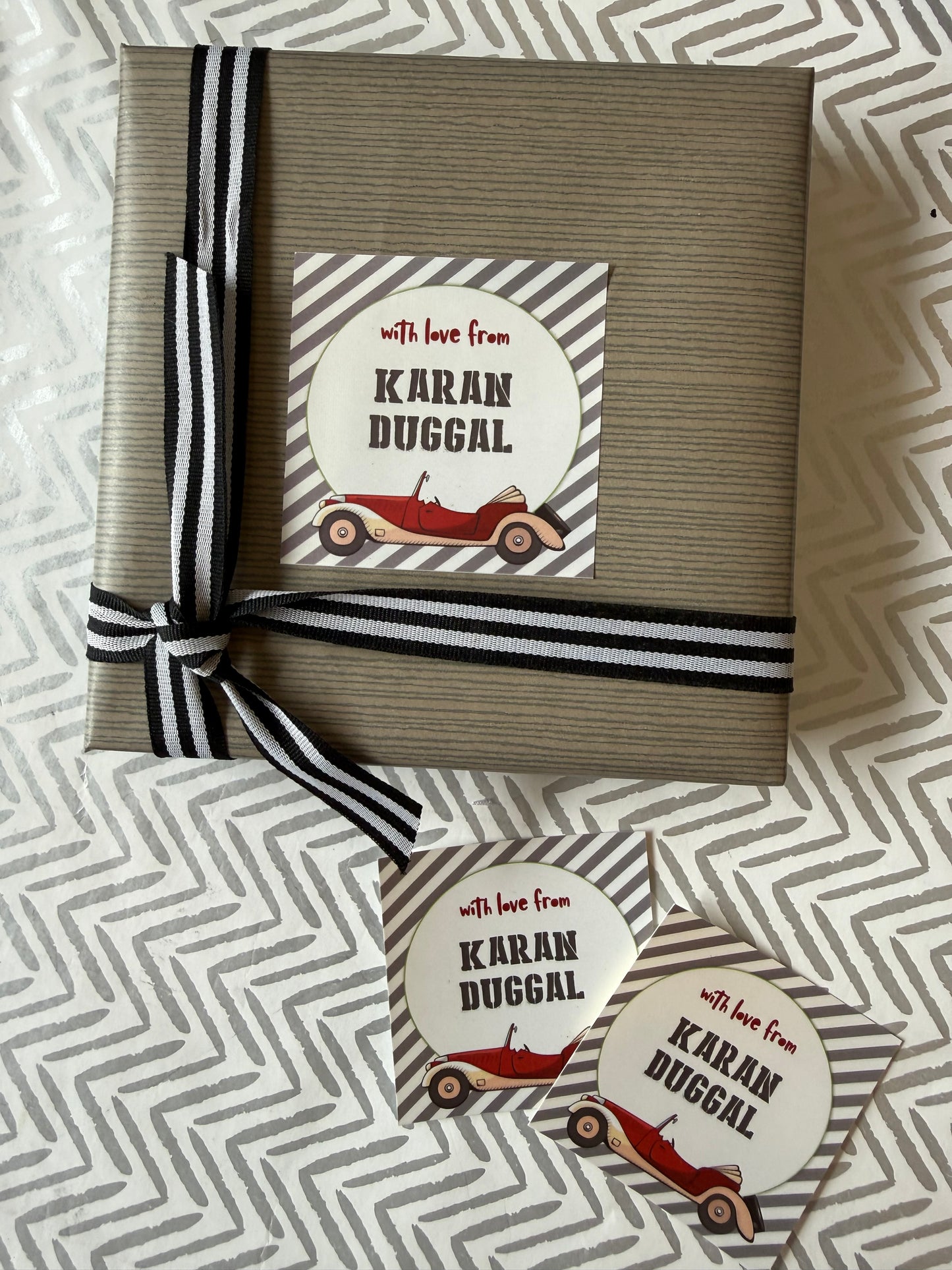 A wrapped gift box and two matching round stickers on a patterned fabric background. The box is wrapped in taupe paper with a black-and-white striped ribbon, and the round sticker reads 'with love from Karan Duggal' with a red vintage car illustration.