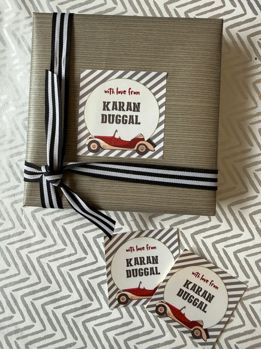 A wrapped gift box and two matching round stickers on a patterned fabric background. The box is wrapped in taupe paper with a black-and-white striped ribbon, and the round sticker reads 'with love from Karan Duggal' with a red vintage car illustration.