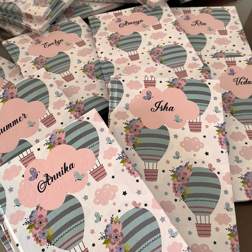 A collection of personalized notebooks with a balloon and star pattern, featuring names such as Anika, Isha, and Vedika on the covers.
