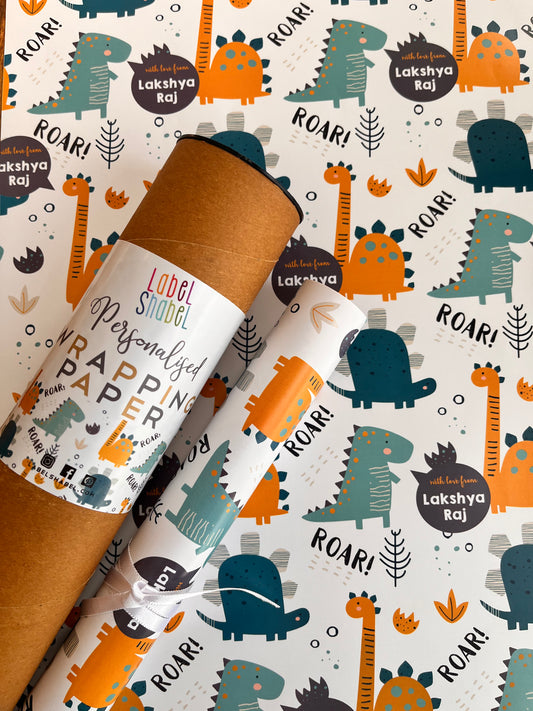 Two rolls of wrapping paper with a playful dinosaur and sound-themed pattern, displayed diagonally on top of a matching roll of brown kraft paper. The pattern features cartoon dinosaurs in teal and orange, with text like 'ROAR!'