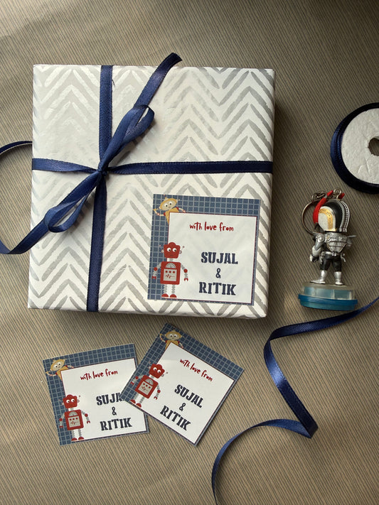 A wrapped gift box with a navy blue ribbon, featuring a personalized gift tag that reads 'with love from SJJAL & RITIK' with cartoon robot illustrations. Two matching tags are placed beside it on a gray textured surface, with a roll of navy ribbon and a small decorative object nearby.