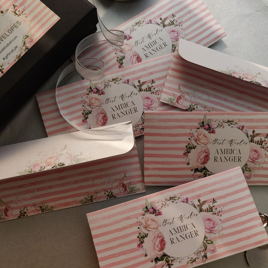 A set of pink and white striped envelope-style stationery pieces with floral rose prints and circular labels reading 'Best Wishes AMBICA RANGER,' arranged on a neutral surface.