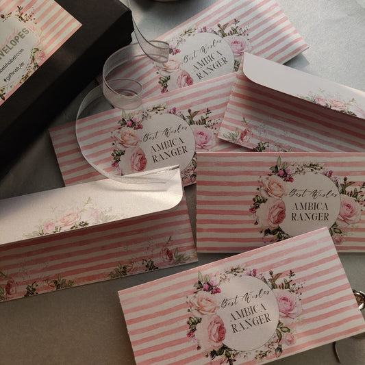A set of pink and white striped envelope-style stationery pieces with floral rose prints and circular labels reading 'Best Wishes AMBICA RANGER,' arranged on a neutral surface.
