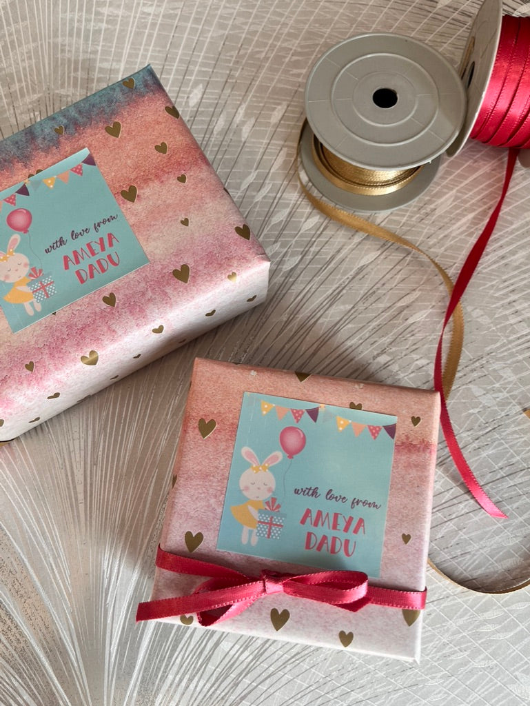Two wrapped gift boxes with pastel pink and white patterned paper, heart prints, and light blue labels reading 'with love from AMEYA DADU,' styled on a light textured surface with a spool of ribbon nearby.