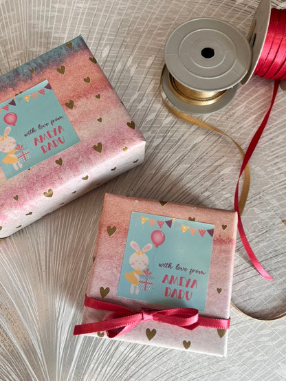 Two wrapped gift boxes with pastel pink and white patterned paper, heart prints, and light blue labels reading 'with love from AMEYA DADU,' styled on a light textured surface with a spool of ribbon nearby.