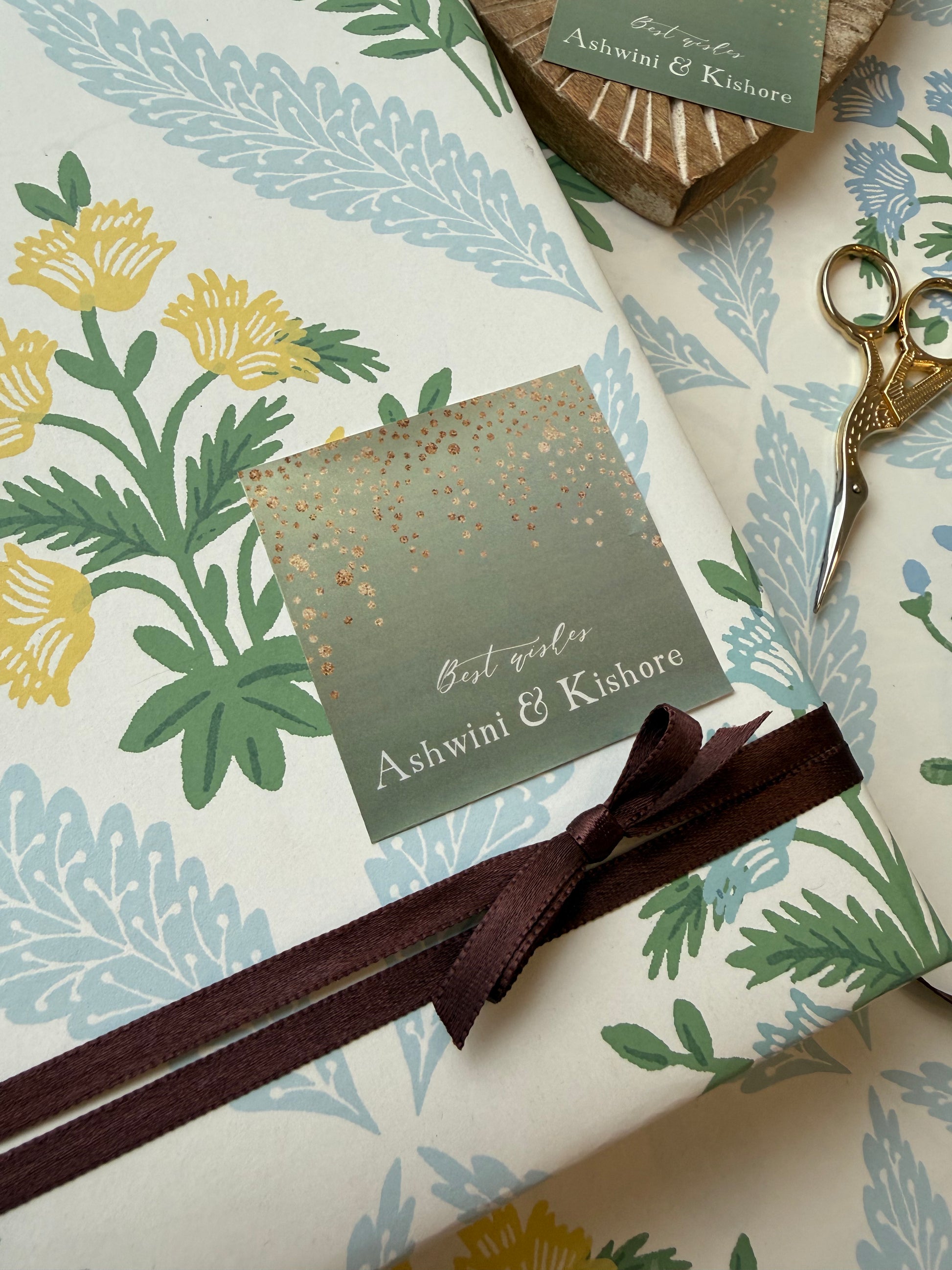 A square sage green card with gold confetti and the names 'Ashwini & Kishore' is attached, with matching scissors and a small tag visible in the background.