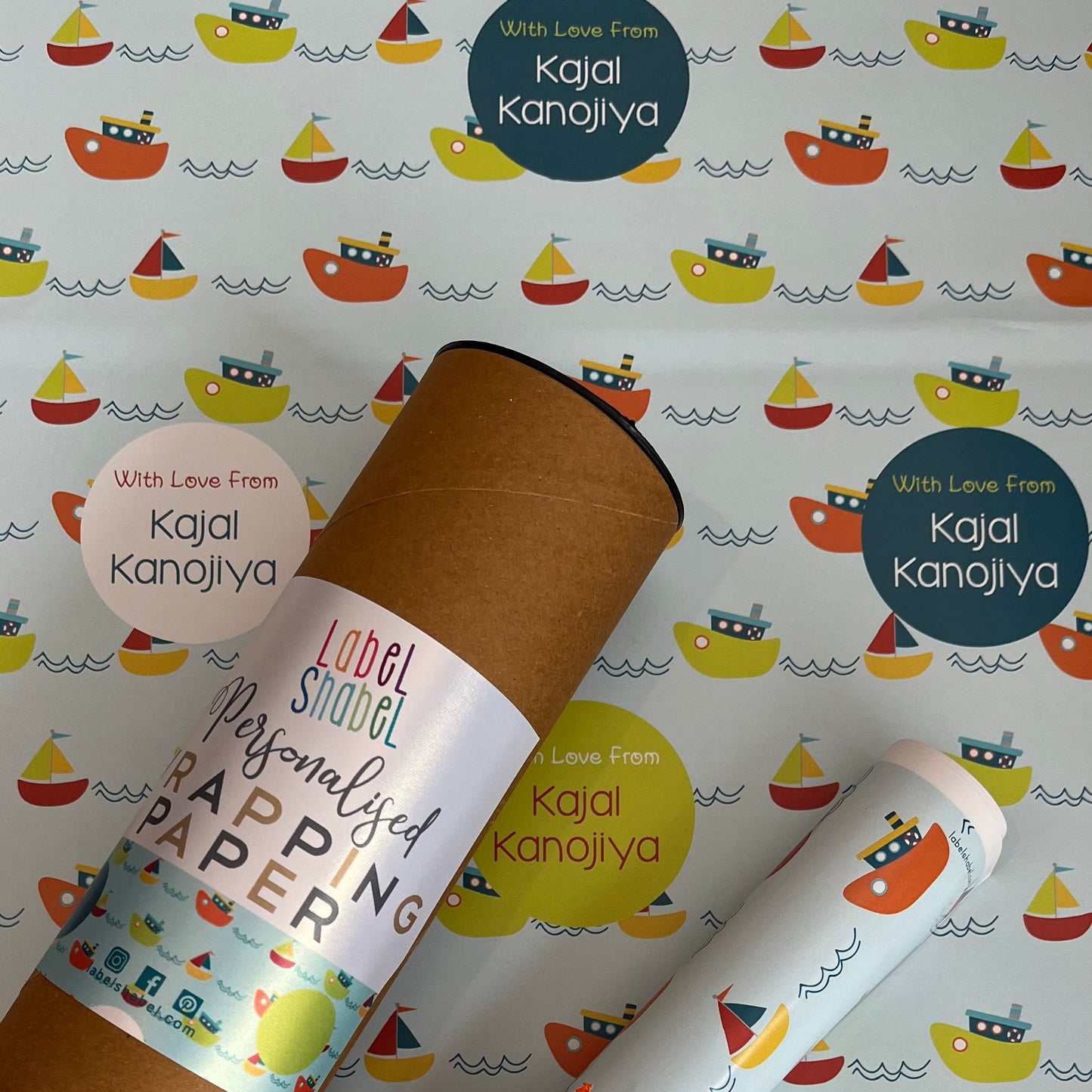 Personalized wrapping paper set featuring a brown kraft cardboard tube and rolled sheets printed with a colorful sailboat pattern, tied with an orange ribbon, on a matching patterned background. Visible text includes 'Kajal Kanojiya' and 'Love From Kajal Kanojiya'.