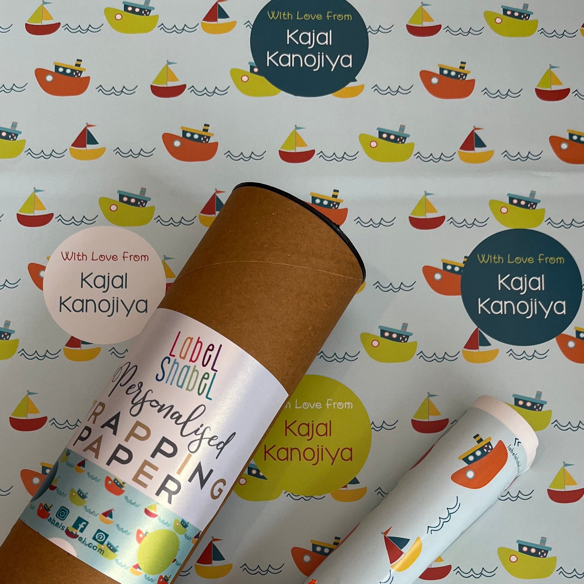 Personalized wrapping paper set featuring a brown kraft cardboard tube and rolled sheets printed with a colorful sailboat pattern, tied with an orange ribbon, on a matching patterned background. Visible text includes 'Kajal Kanojiya' and 'Love From Kajal Kanojiya'.