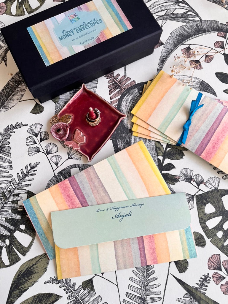 A styled flat-lay of personalized money envelopes with pastel watercolor stripes, a black gift box labeled 'Personalised Money Envelopes', a small maroon jewelry dish, and a tropical leaf patterned surface.