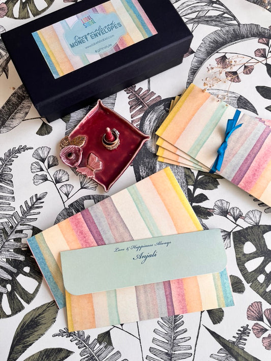 A styled flat-lay of personalized money envelopes with pastel watercolor stripes, a black gift box labeled 'Personalised Money Envelopes', a small maroon jewelry dish, and a tropical leaf patterned surface.