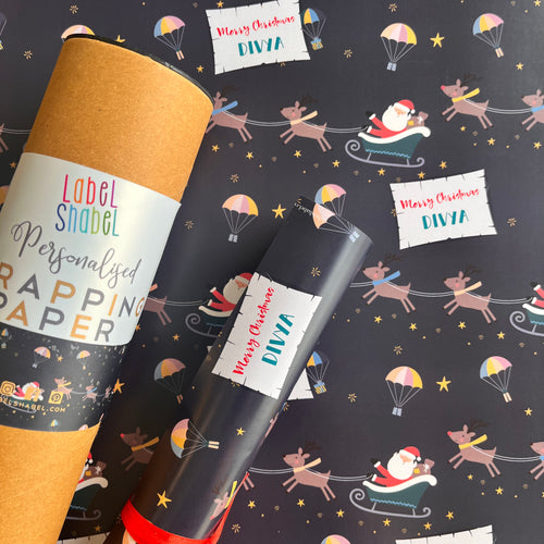 A roll of personalized wrapping paper with a Christmas theme, featuring Santa's sleigh, reindeer, and festive patterns. The paper is wrapped around a cylindrical object, which appears to be a label or tag, and is placed against a similar patterned background.