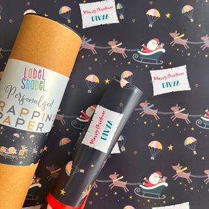 A roll of personalized wrapping paper with a Christmas theme, featuring Santa's sleigh, reindeer, and festive patterns. The paper is wrapped around a cylindrical object, which appears to be a label or tag, and is placed against a similar patterned background.