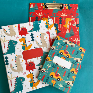 A set of school supplies with dinosaur-themed covers, including a notebook, a clipboard, and a folder, displayed on a teal background.