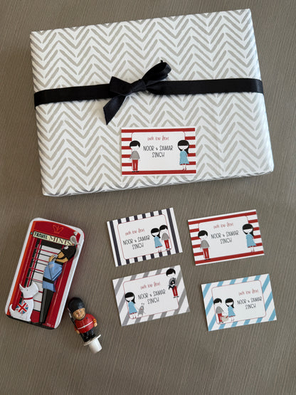 A gift-wrapped box and matching printed cards laid out on a neutral surface. The box is wrapped in a white and gray chevron pattern with a black ribbon, and the printed pieces show red-and-white striped invitation cards, smaller matching cards, a red telephone booth illustration, and a small toy figure.