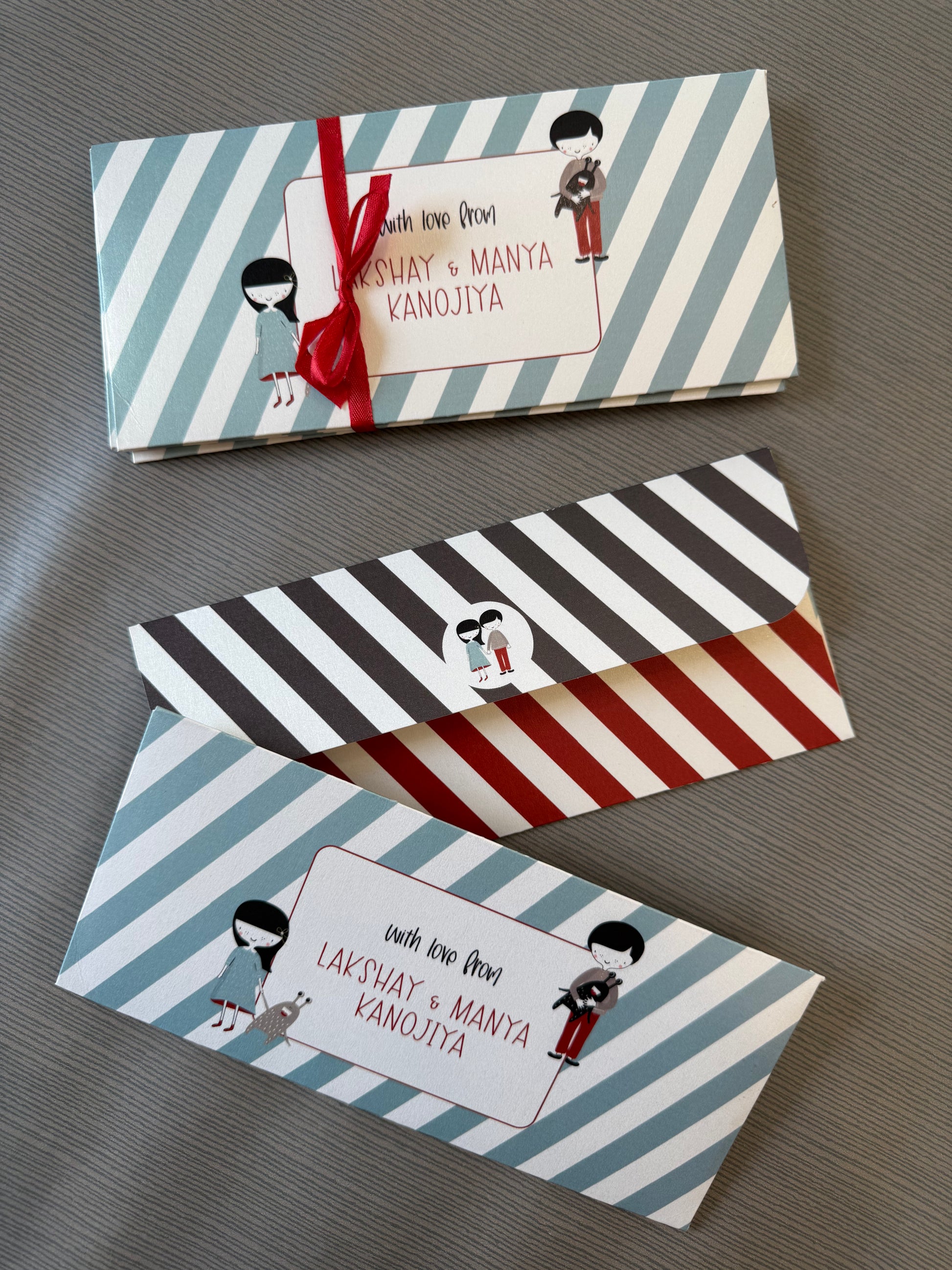 Three personalized paper gift envelopes with striped patterns and cartoon illustrations, arranged diagonally on a gray textured surface. The top envelope has light blue and white stripes with a red ribbon, the middle has black, white, and red stripes, and the bottom has light blue, white, and red stripes.