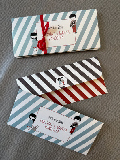 Three personalized paper gift envelopes with striped patterns and cartoon illustrations, arranged diagonally on a gray textured surface. The top envelope has light blue and white stripes with a red ribbon, the middle has black, white, and red stripes, and the bottom has light blue, white, and red stripes.