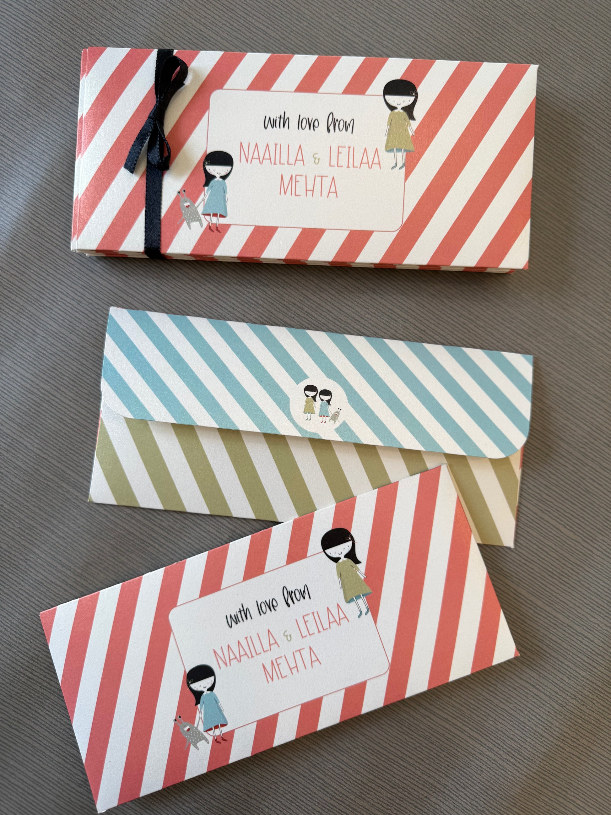 Three striped gift envelopes or card holders arranged on a gray textured surface. The top item has coral and white diagonal stripes with a black ribbon and a white label reading 'with love from NAAILLA & LEILAA MEHTA.