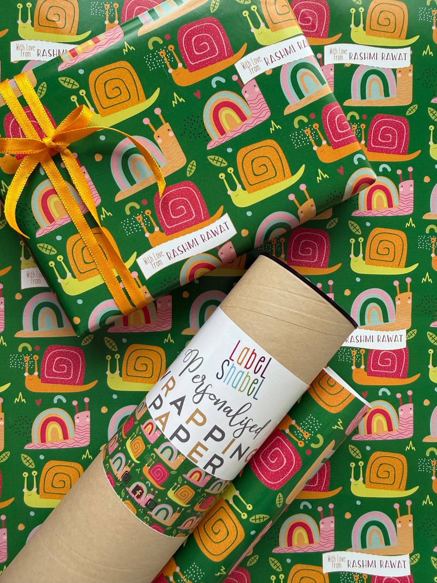 A roll of wrapping paper and a wrapped gift box with a matching patterned wrap, featuring a colorful retro snail design on a green background. The gift is tied with a yellow ribbon, 