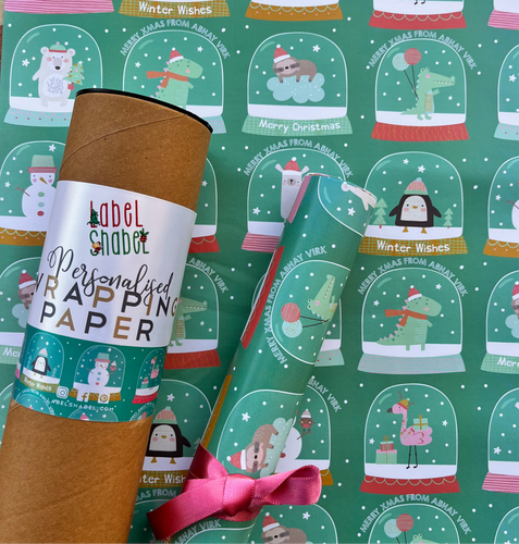 A roll of personalized wrapping paper with Christmas-themed designs including snow globes, mistletoe, and cute animal illustrations, placed next to a label that reads 'Personalised Wrapping Paper'.