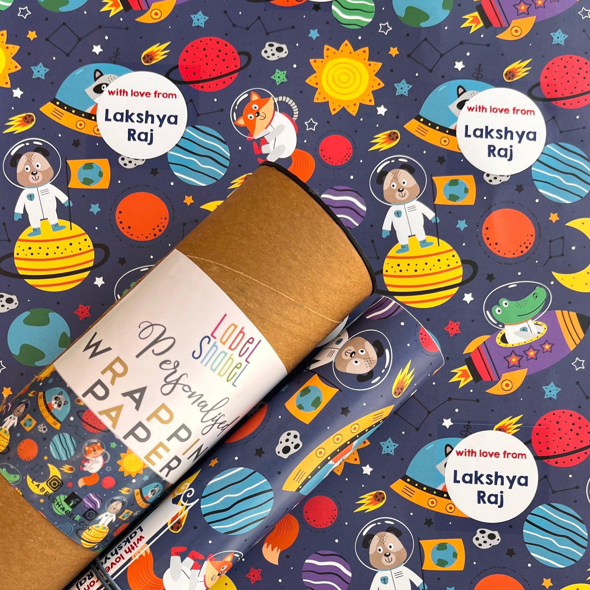 A rolled kraft paper gift wrap or mailing tube placed diagonally over a matching printed sheet featuring a colorful space-themed pattern with planets, astronauts, rockets, and the names Lakshya Raj.