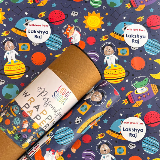 A rolled kraft paper gift wrap or mailing tube placed diagonally over a matching printed sheet featuring a colorful space-themed pattern with planets, astronauts, rockets, and the names Lakshya Raj.