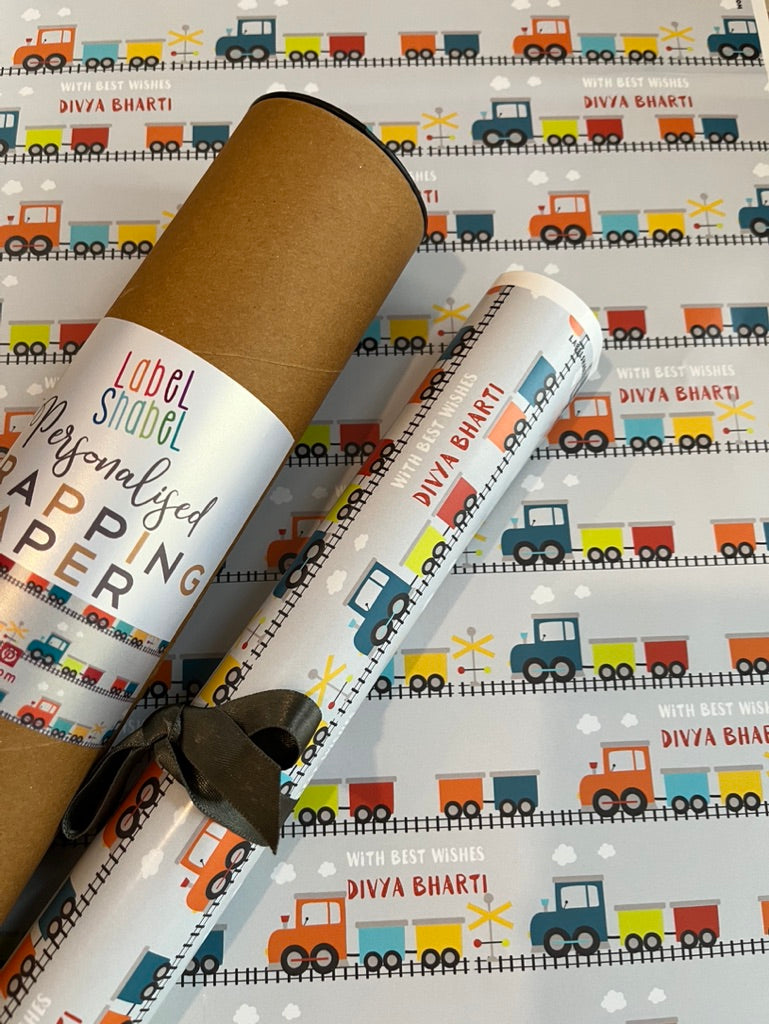 Personalised baby shower wrapping paper featuring a colorful cartoon train pattern on a white background, shown partially rolled with a brown cardboard mailing tube