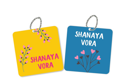 Two square metal bag tags with floral designs and customizable names, one in yellow with pink flowers and the other in blue with gray flowers.