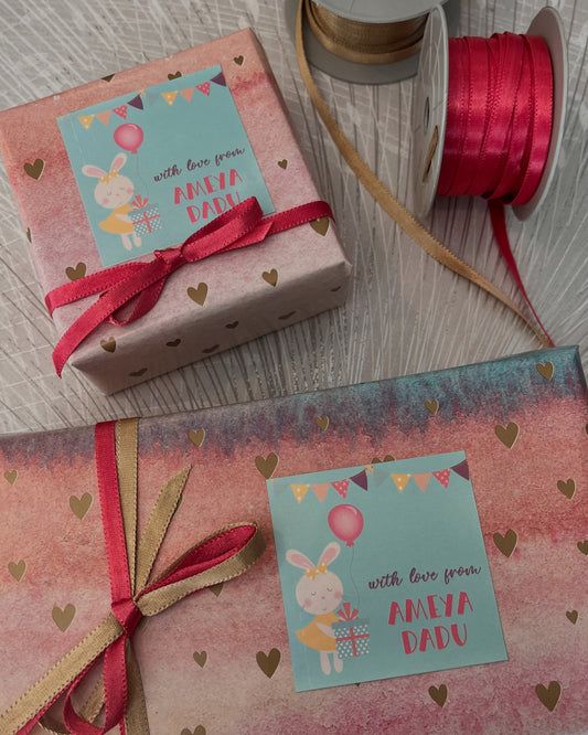 A styled flat-lay of a pink and teal gift box and matching envelope with gold heart patterns, personalized with a cartoon bunny card reading 'with love from AMEYA DADU', tied with red and gold ribbons, with a spool of pink ribbon on a light wood background.