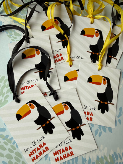 Several white gift tags with toucan illustrations and hanging ribbons are arranged on a light patterned surface. The tags include the text 'love & luck' and 'NITARA MAHAR'.