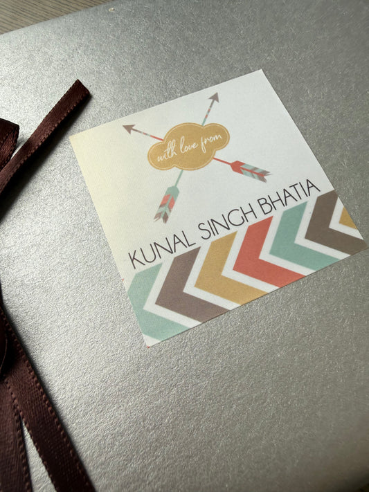 A square printed card or label with the text 'KUNAL SINH BHAIYA' and decorative chevron stripes, placed on a metallic silver surface with a dark brown ribbon partially visible at the bottom.