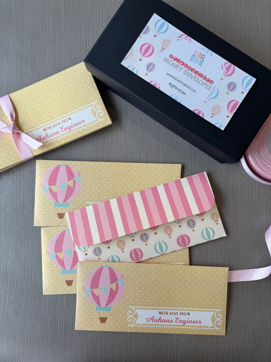 A styled flat-lay of personalized gift envelopes, featuring a black gift box with a hot air balloon patterned label, several gold polka-dot envelopes with pink ribbons and labels, and two bookmarks with pink and white stripes and hot air balloon prints, all arranged on a gray textured surface.