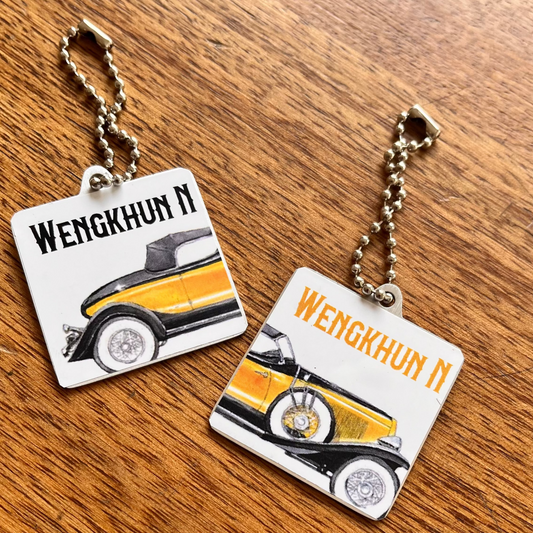 Two small square metal keychains with chain loops, shown on a wooden tabletop. Each keychain has a white square charm featuring a vintage car illustration and the text 'WENGKHUN N' in orange/yellow, with one charm showing a black-and-yellow car and the other showing a yellow-and-black car.