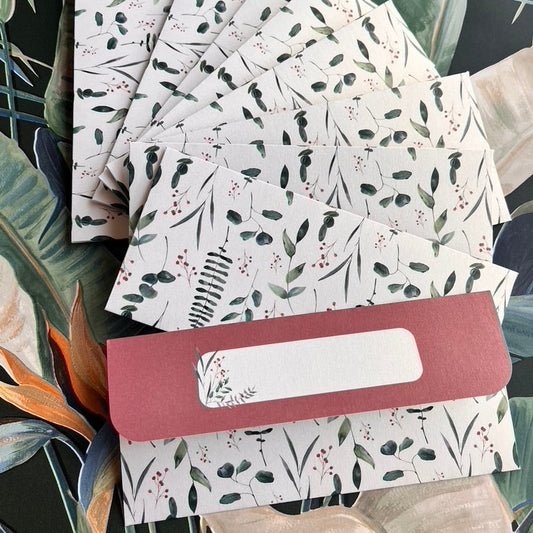A fan arrangement of white paper envelopes printed with a green botanical leaf pattern, held together by a dusty rose paper band with a blank white label slot, set against a tropical leaf background.