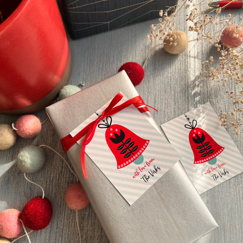 Ribbon tags with 'Holiday Bells' design, attached to a gift wrapped in a red ribbon, presented on a table with decoration items and a cup in the background.