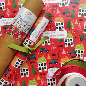 A roll of personalized wrapping paper with a Christmas theme, including houses with Christmas decorations, trees, and festive text.