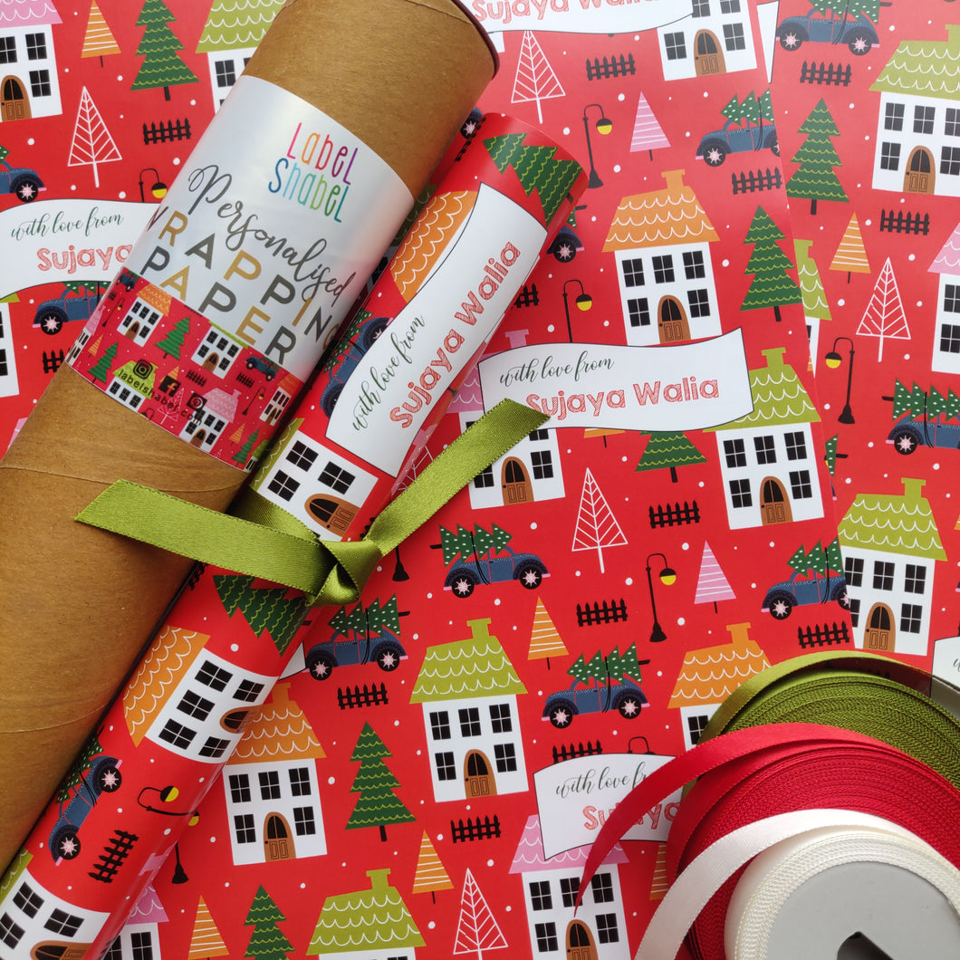 A roll of personalized wrapping paper with a Christmas theme, including houses with Christmas decorations, trees, and festive text.