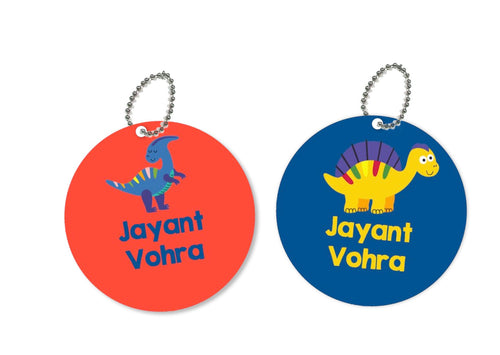 Two round metal bag tags with dinosaur illustrations and names 'Jayant' and 'Vohra' on them, one in blue and the other in orange.
