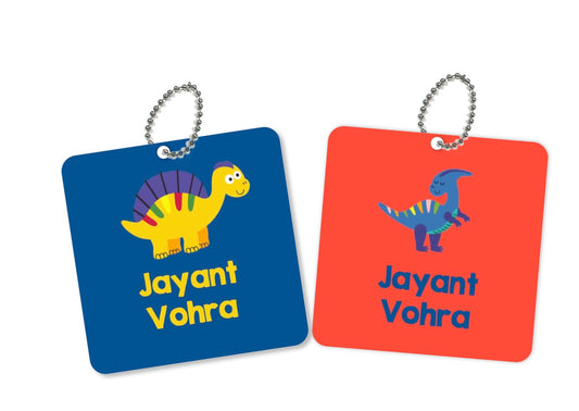 Two square metal bag tags with dinosaur illustrations and names 'Jayant' and 'Vohra' on them, one in blue and the other in orange.